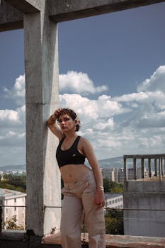 Confident woman in sportswear leans against a concrete post on a rooftop under a bright blue sky.