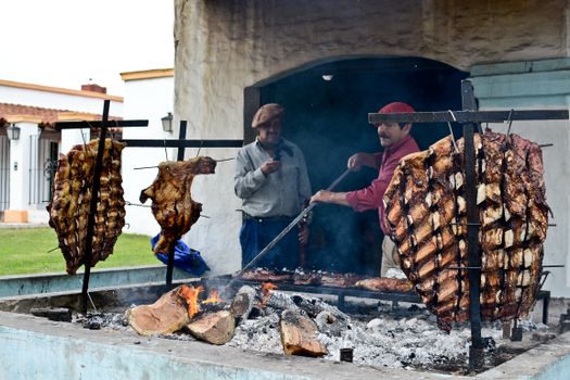 Argentine men preparing traditional asado with meats grilling over open flame outdoors.