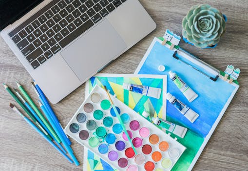 Top-down view of a creative workspace featuring watercolor paints, brushes, and a laptop.
