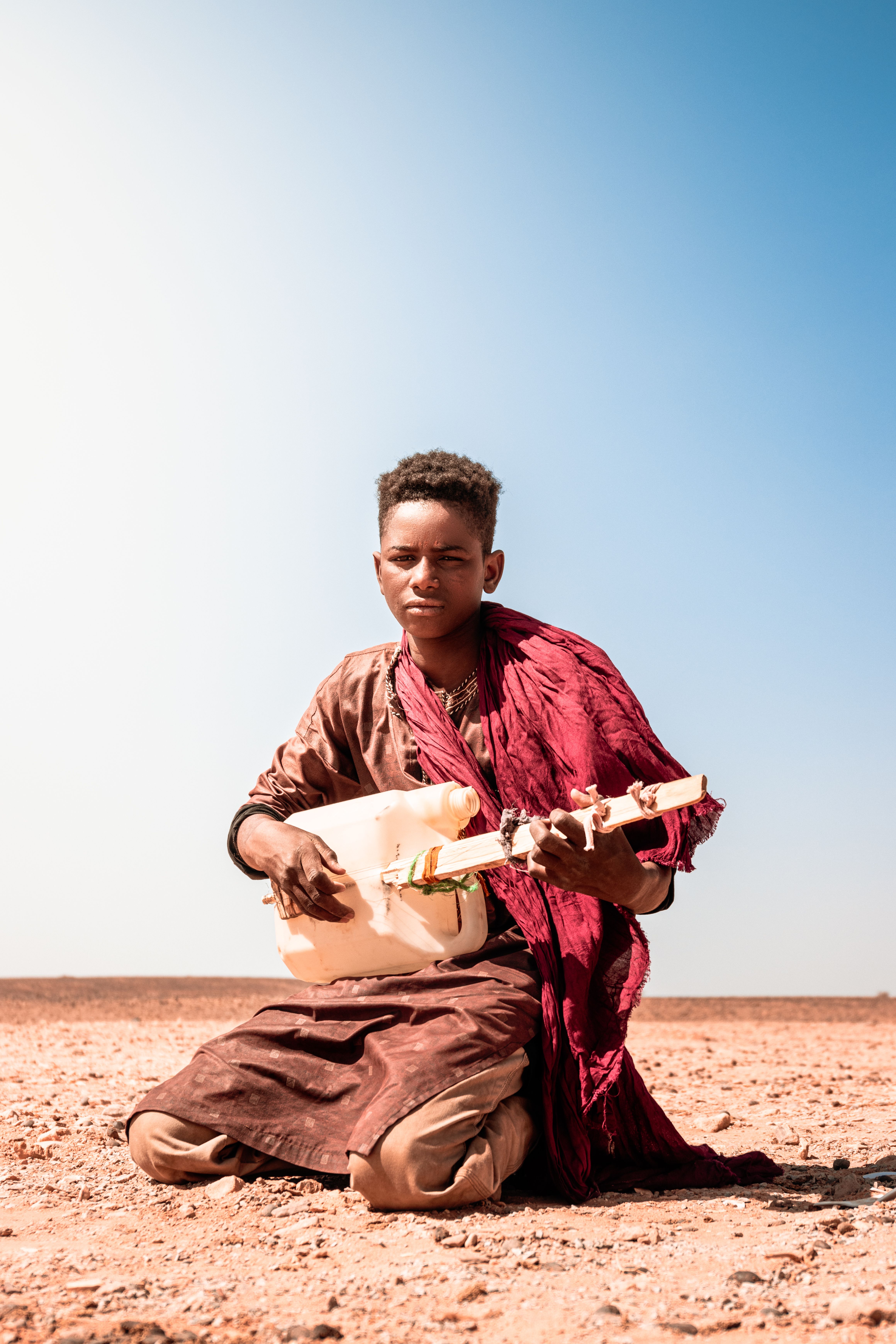 A teenage boy plays a homemade guitar while sitting in an arid desert setting under a clear blue sky.