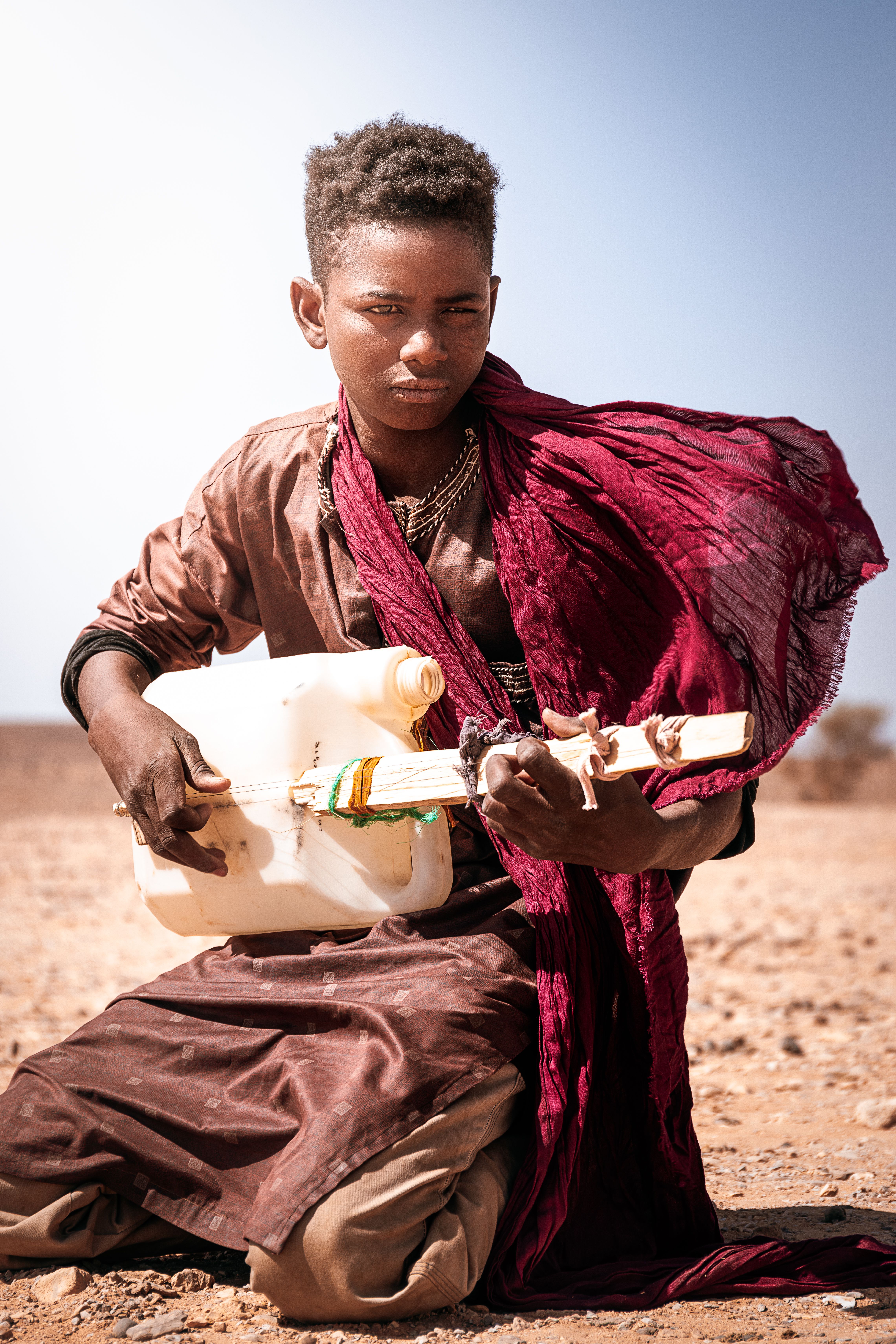 Young boy plays a makeshift guitar in a desert setting, a red scarf blowing in the wind.