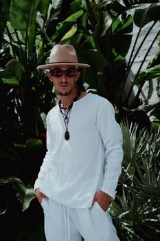 Fashionable man in white attire posing amidst lush tropical plants, wearing a hat and sunglasses.