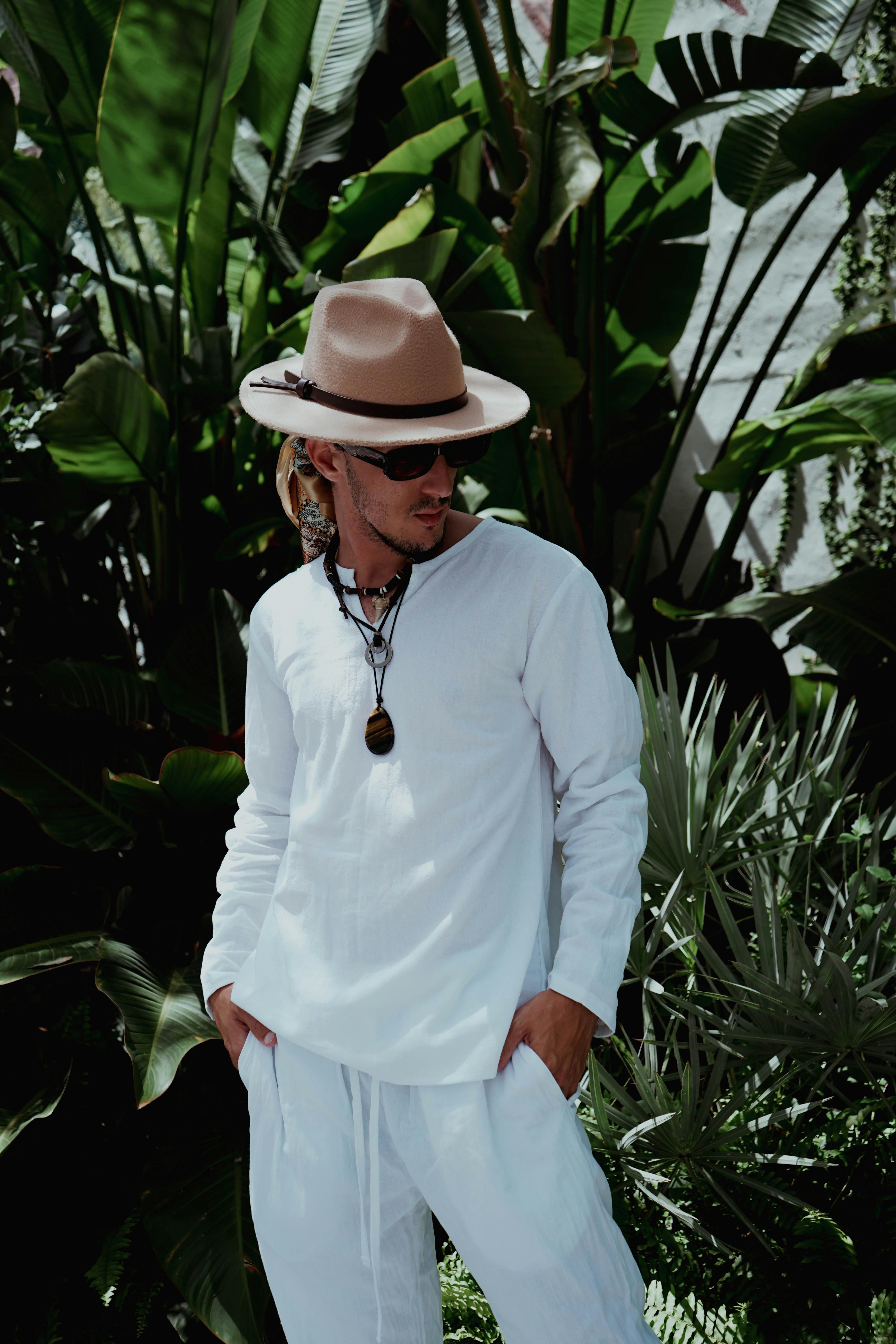 Young man in white clothing, sunglasses, and hat posing amidst lush greenery.