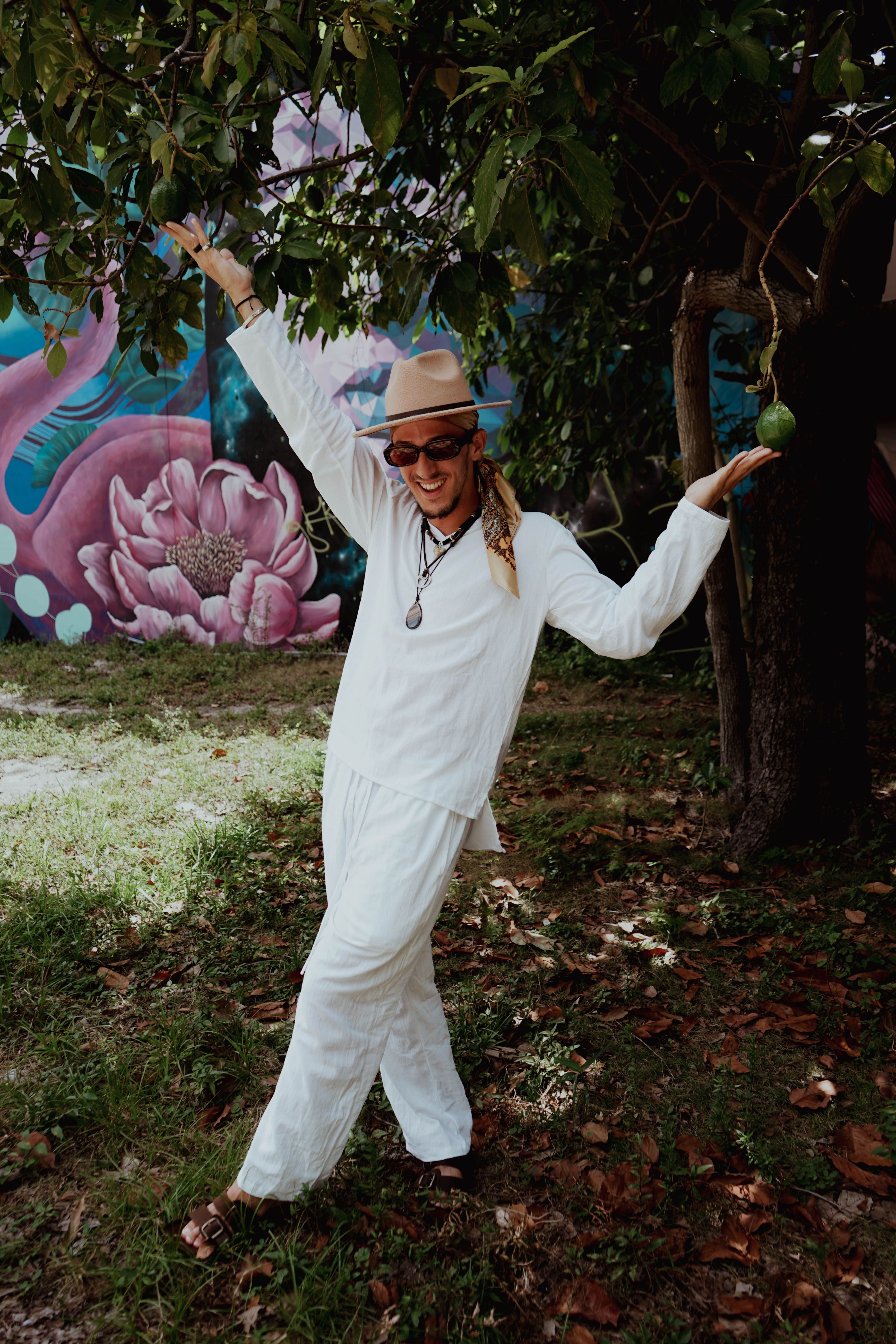 A fashionable man in a white outfit joyfully poses under a tree beside a colorful mural.