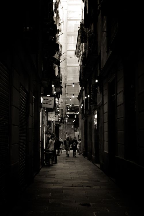 Free Black and white photo of a shadowy Barcelona alley with people walking, capturing urban mood. Stock Photo