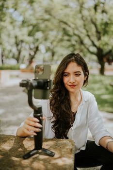 Young woman with long hair filming with a smartphone and gimbal in a sunny outdoor park setting.