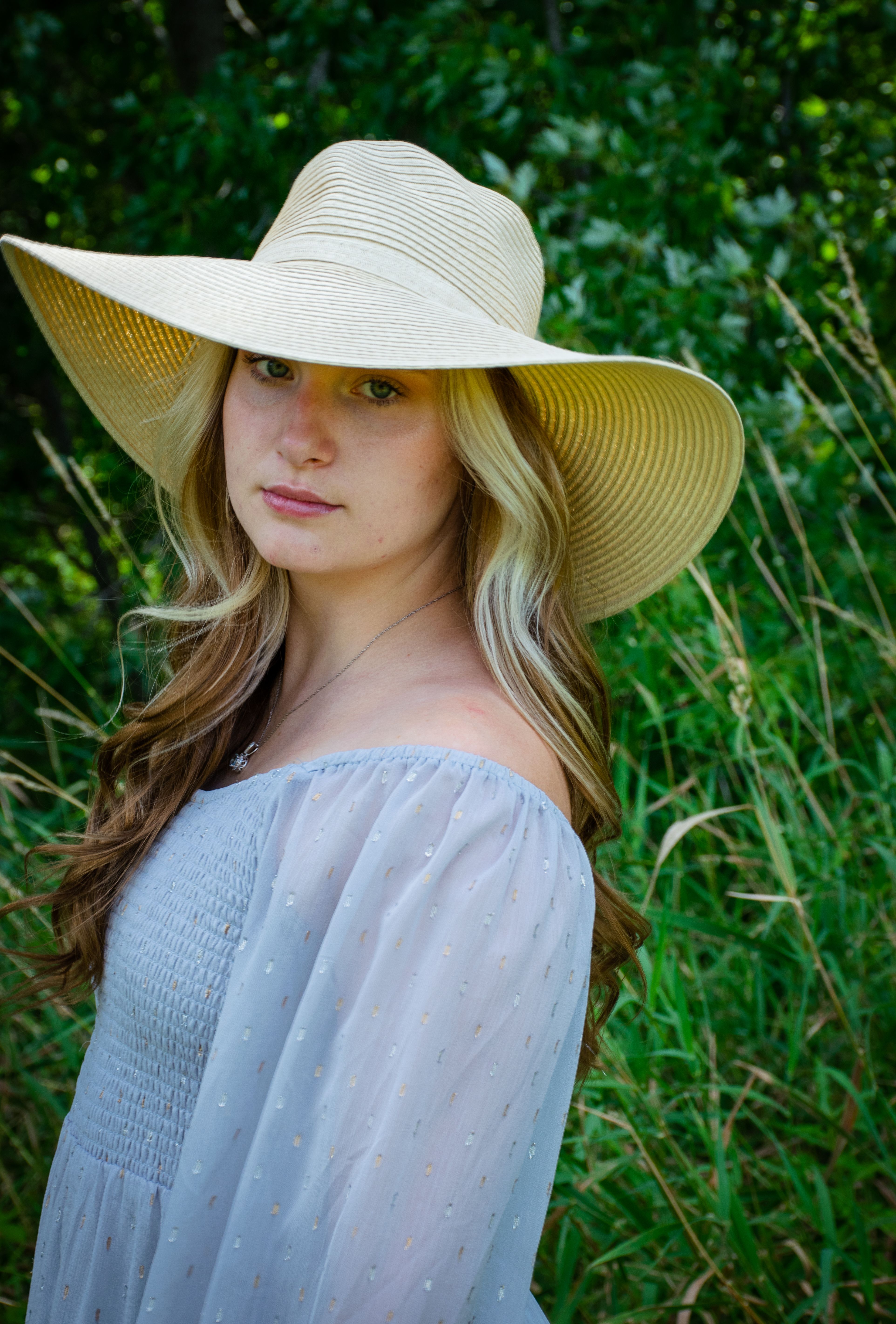 Elegant portrait of a woman in a straw hat posing outdoors against a lush green backdrop.