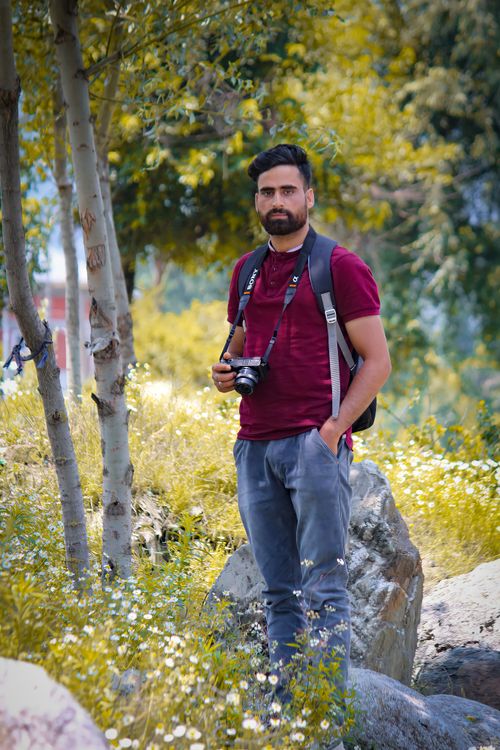Free Man standing with camera amidst rocks and trees, wearing casual outdoor attire. Stock Photo