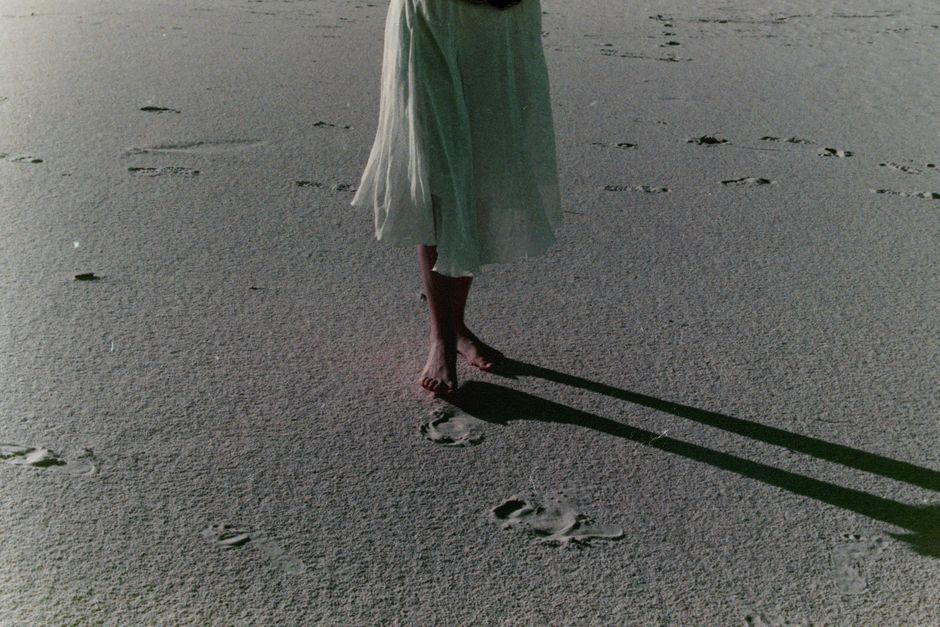 A woman's feet walking barefoot on a beach, leaving footprints in the sand.