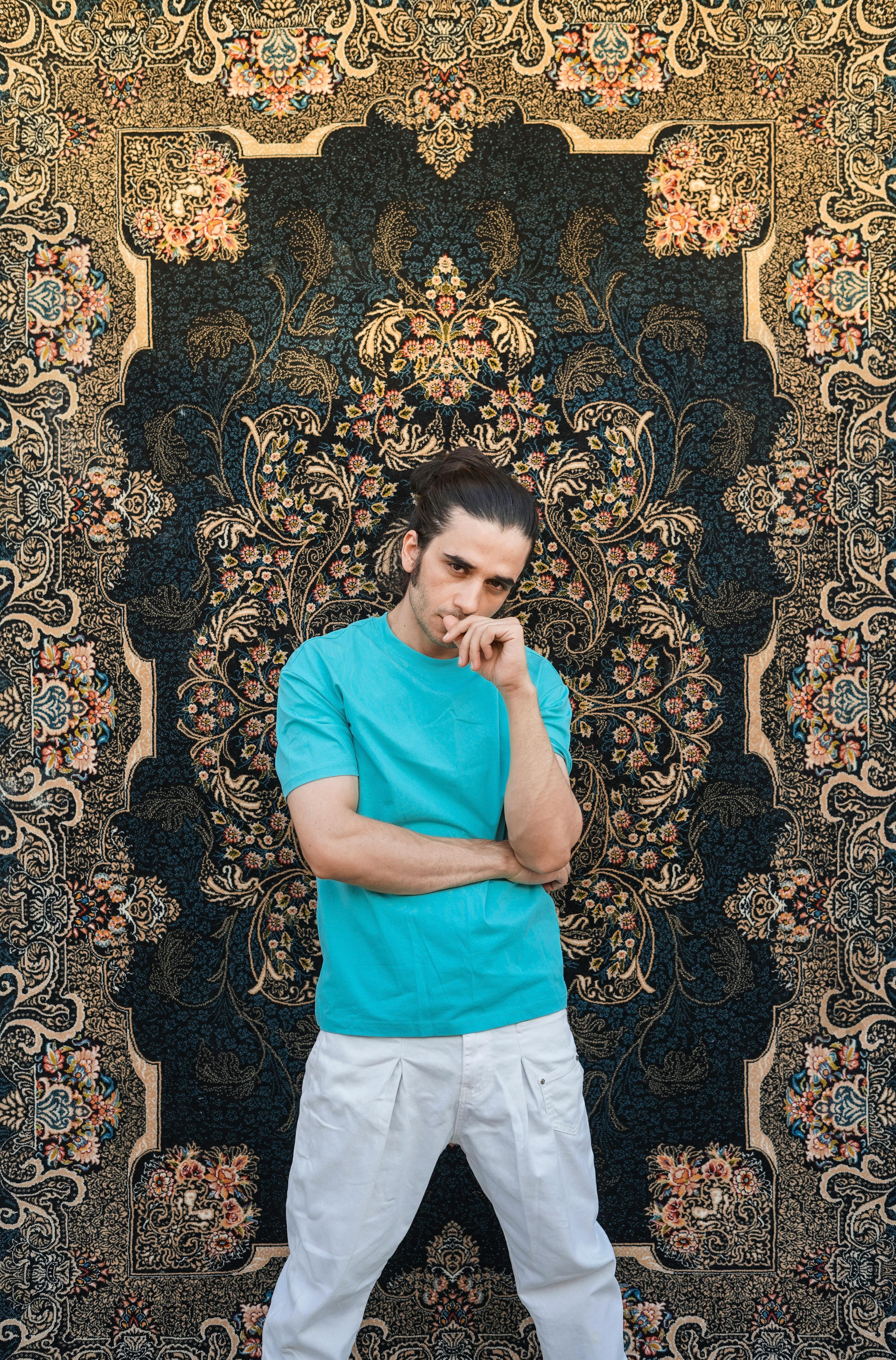 A fashionable man in a turquoise t-shirt and white pants poses confidently against an ornate Persian carpet backdrop.
