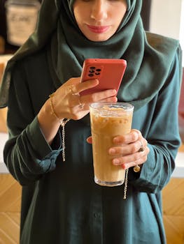 Muslim woman in hijab enjoying iced coffee while using her smartphone indoors.