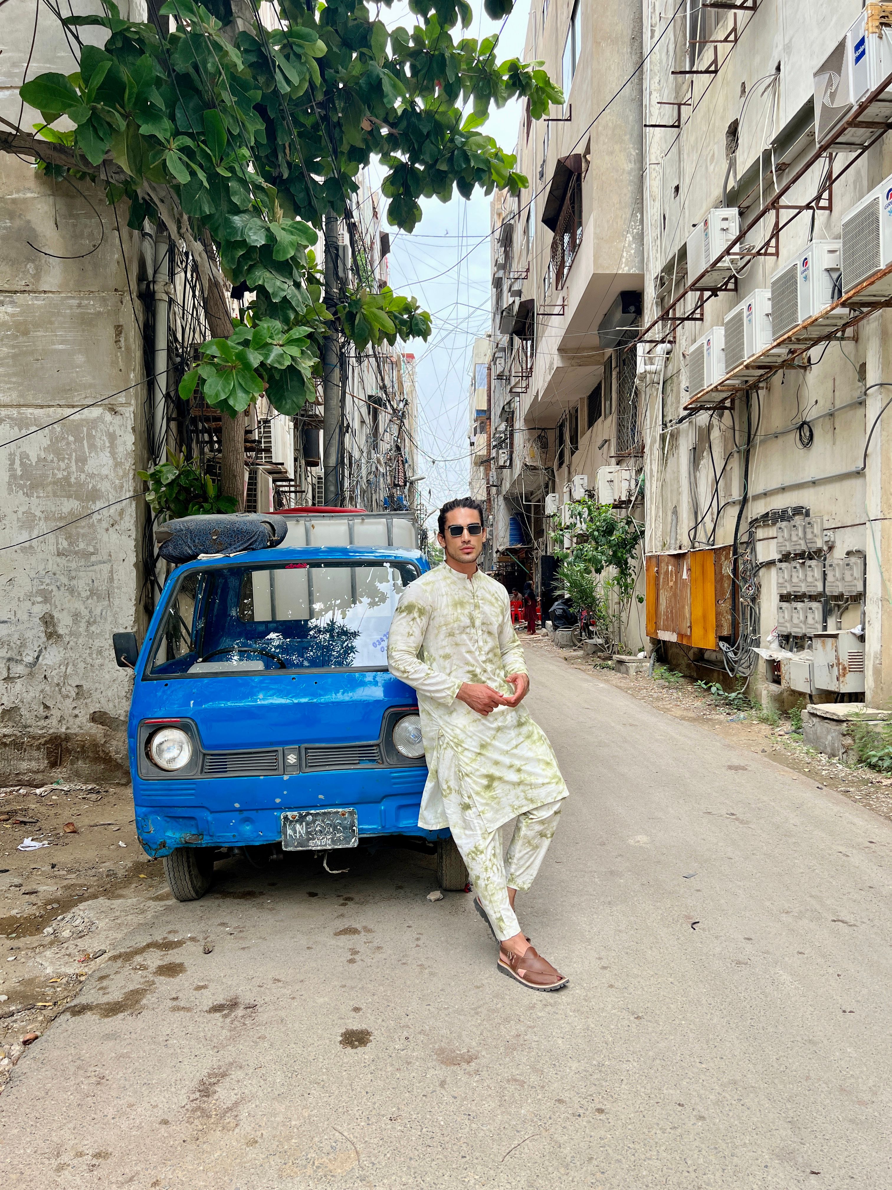 A fashionable man in traditional clothing poses by a blue car in a Karachi alleyway.