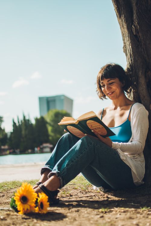 Free A young woman enjoys reading a book by the lakeside under a tree, adding a touch of summer relaxation. Stock Photo