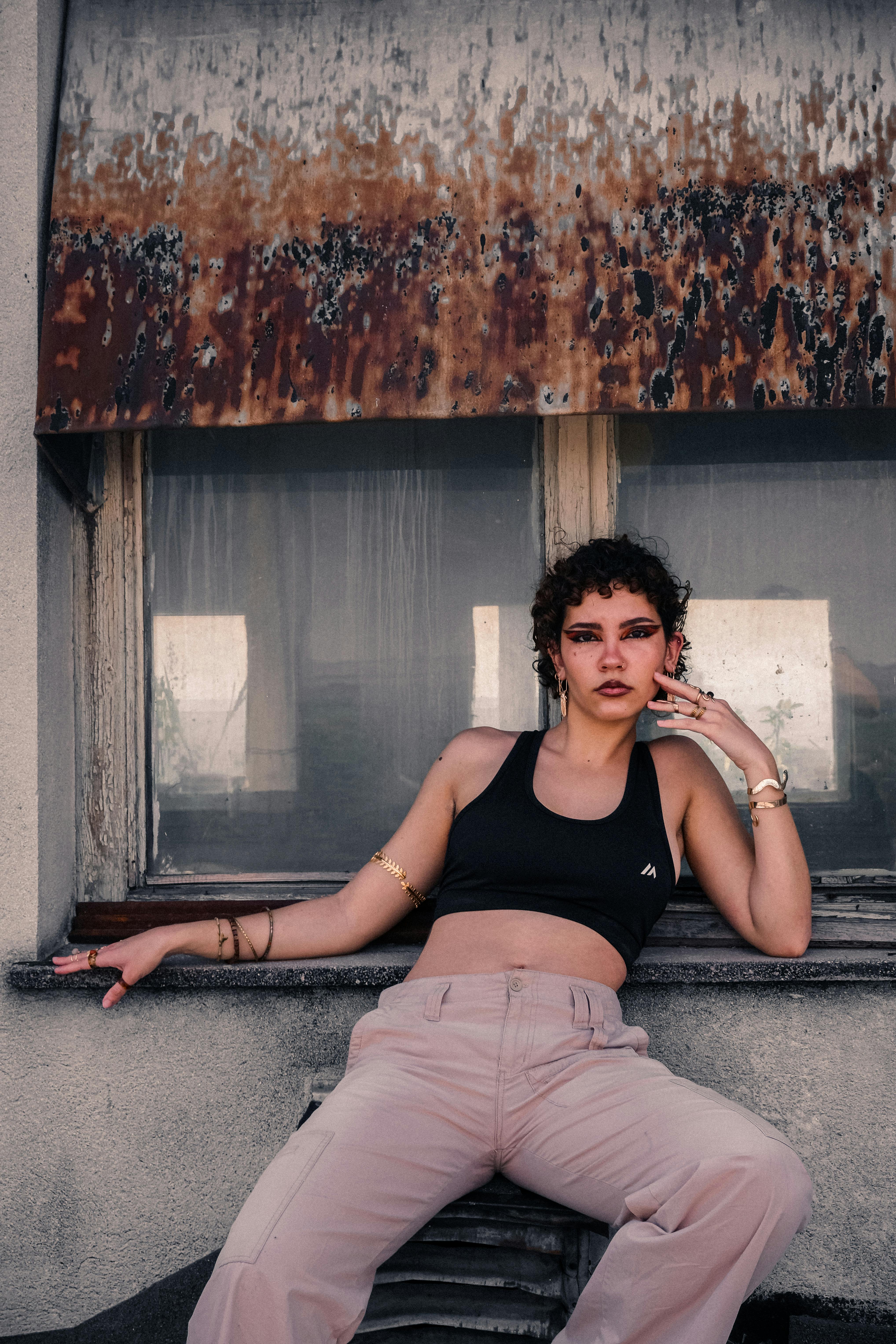 Woman in black tank top posing against a weathered window with rustic backdrop.