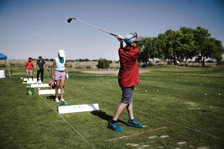 A group of golfers practising swing technique at an outdoor driving range, with an instructor visible in the background observing form and providing guidance.