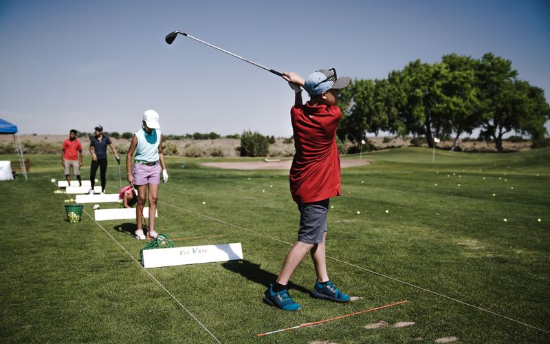 A group of golfers practising swing technique at an outdoor driving range, with an instructor visible in the background observing form and providing guidance.