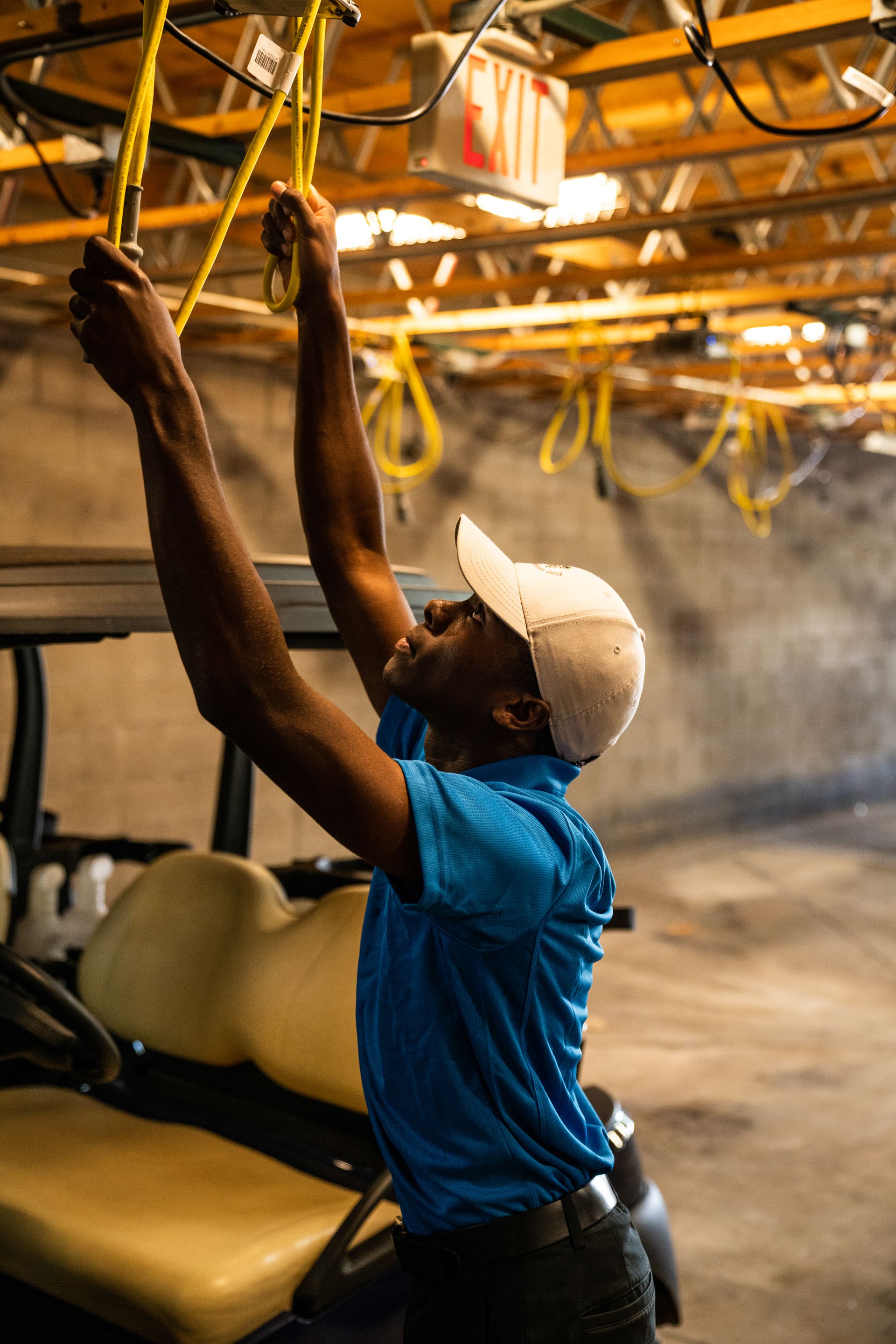 A man wearing a cap works on wiring above a golf cart in a garage.