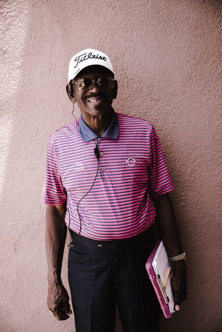 Happy man wearing striped polo and Titleist cap, smiling with earphones. Holding clipboard.