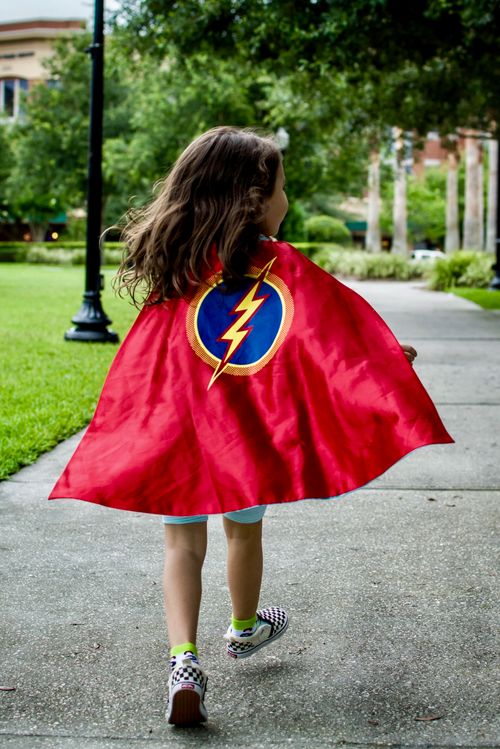 Free A young girl wearing a red superhero cape runs joyfully along a park pathway. Stock Photo