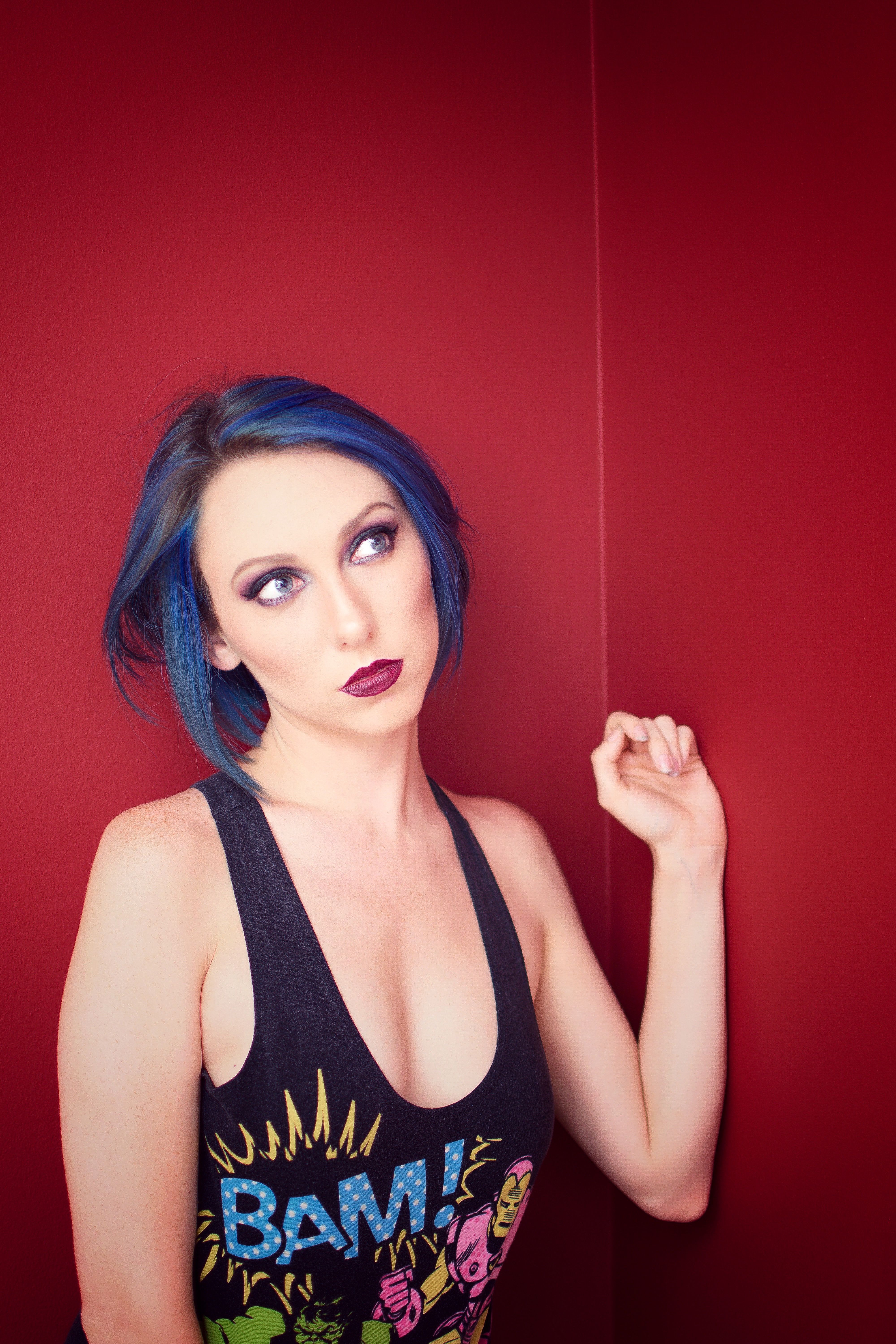 Portrait of a fashionable woman with blue hair and bold makeup posing indoors against a red background.