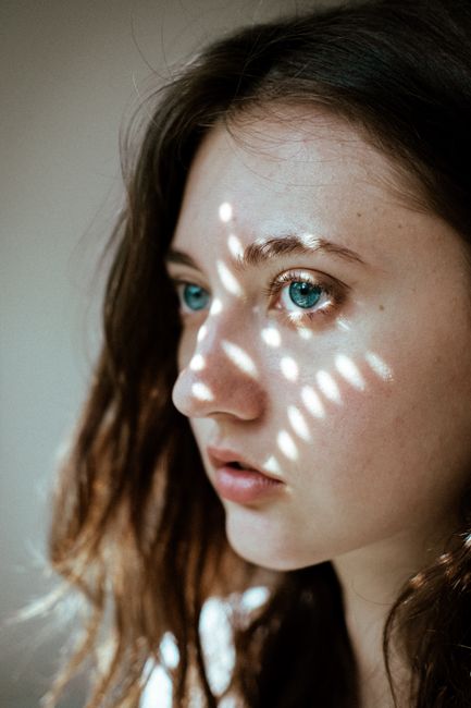 Young woman with striking blue eyes and light patterns on her face, in a thoughtful pose.