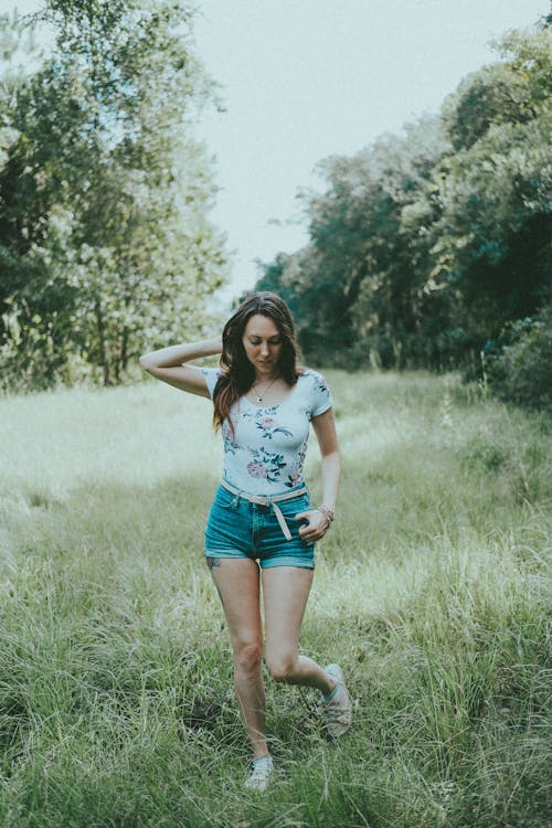 Woman in White Floral Shirt and Denim Shorts Walking on Green Grass Field