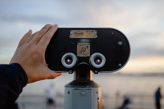 A hand adjusting public viewing binoculars with a scenic sunset in the background.