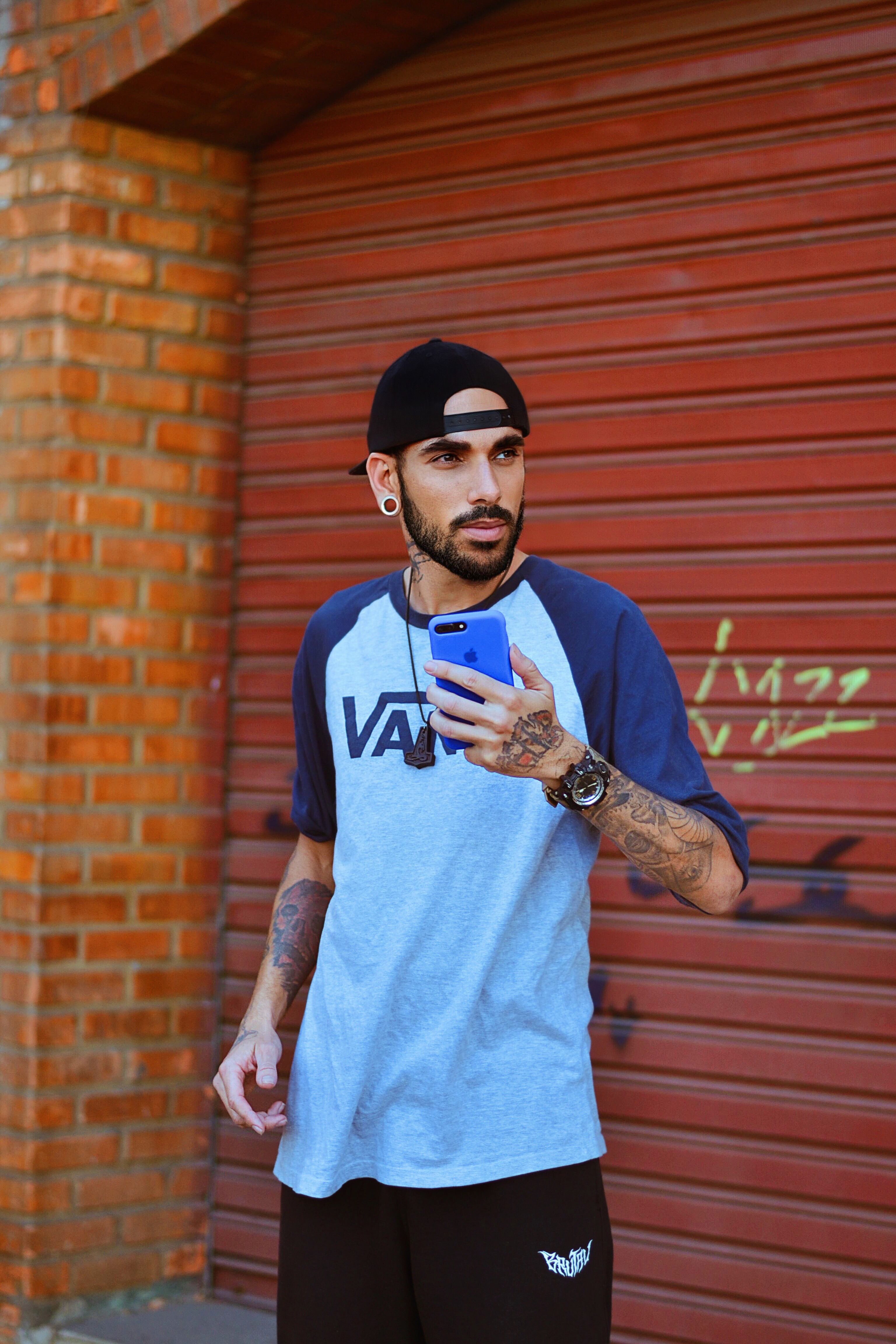 Casual young man with tattoos holding a smartphone against a graffiti background in Sete Lagoas, Brazil.