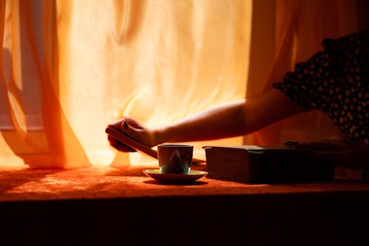 A serene close-up of a hand opening curtains with a teacup and book in warm sunlight.