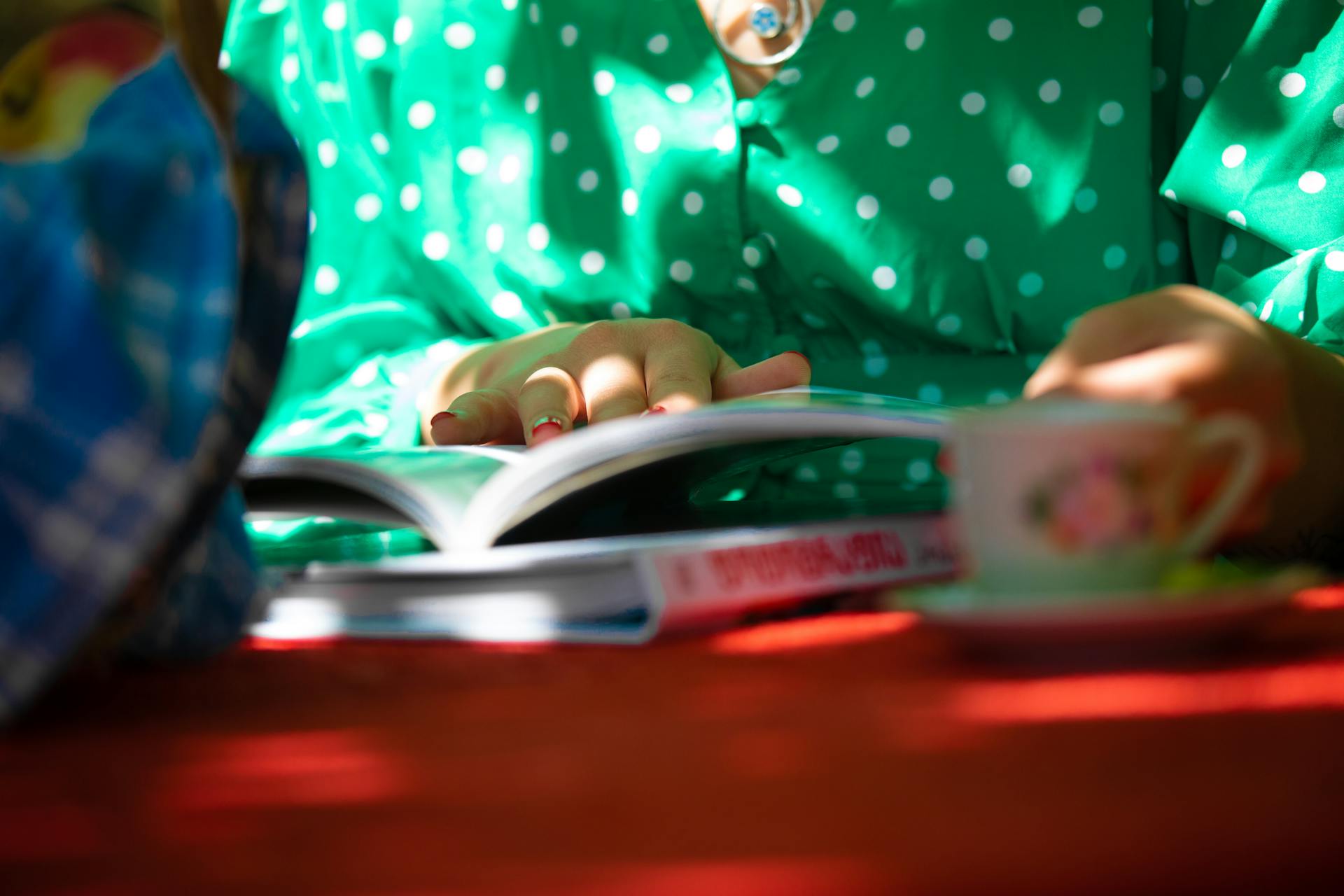 A woman wearing a green polka dot dress reads an open book at a table with a floral teacup.