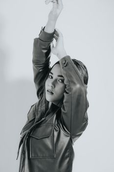 Monochrome portrait of a young woman in leather jacket with arms raised, elegant pose.