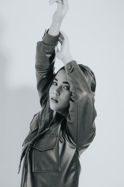 Free Monochrome portrait of a young woman in leather jacket with arms raised, elegant pose. Stock Photo