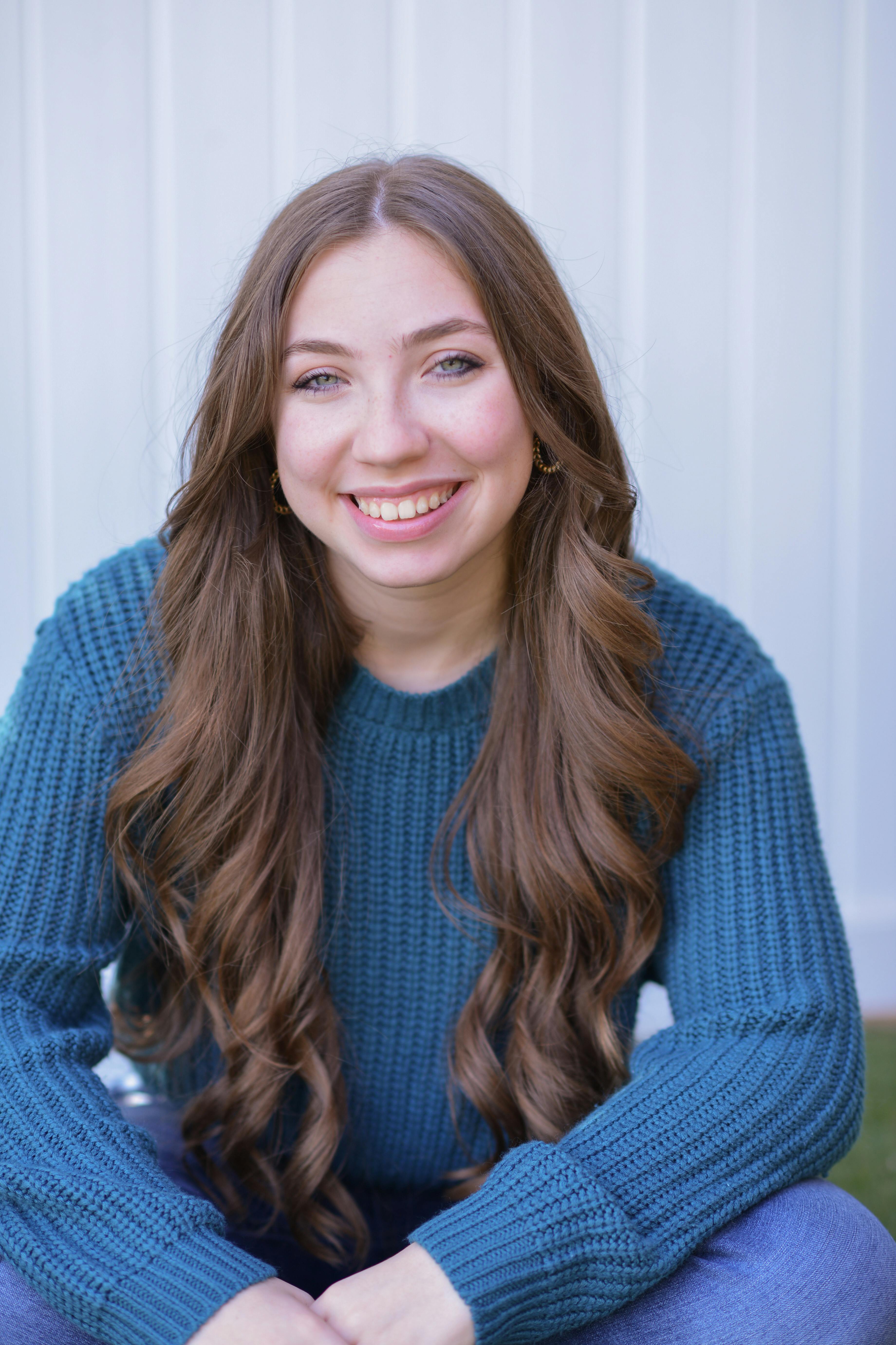 Portrait of a young Caucasian woman with long hair smiling and sitting indoors in a blue sweater.