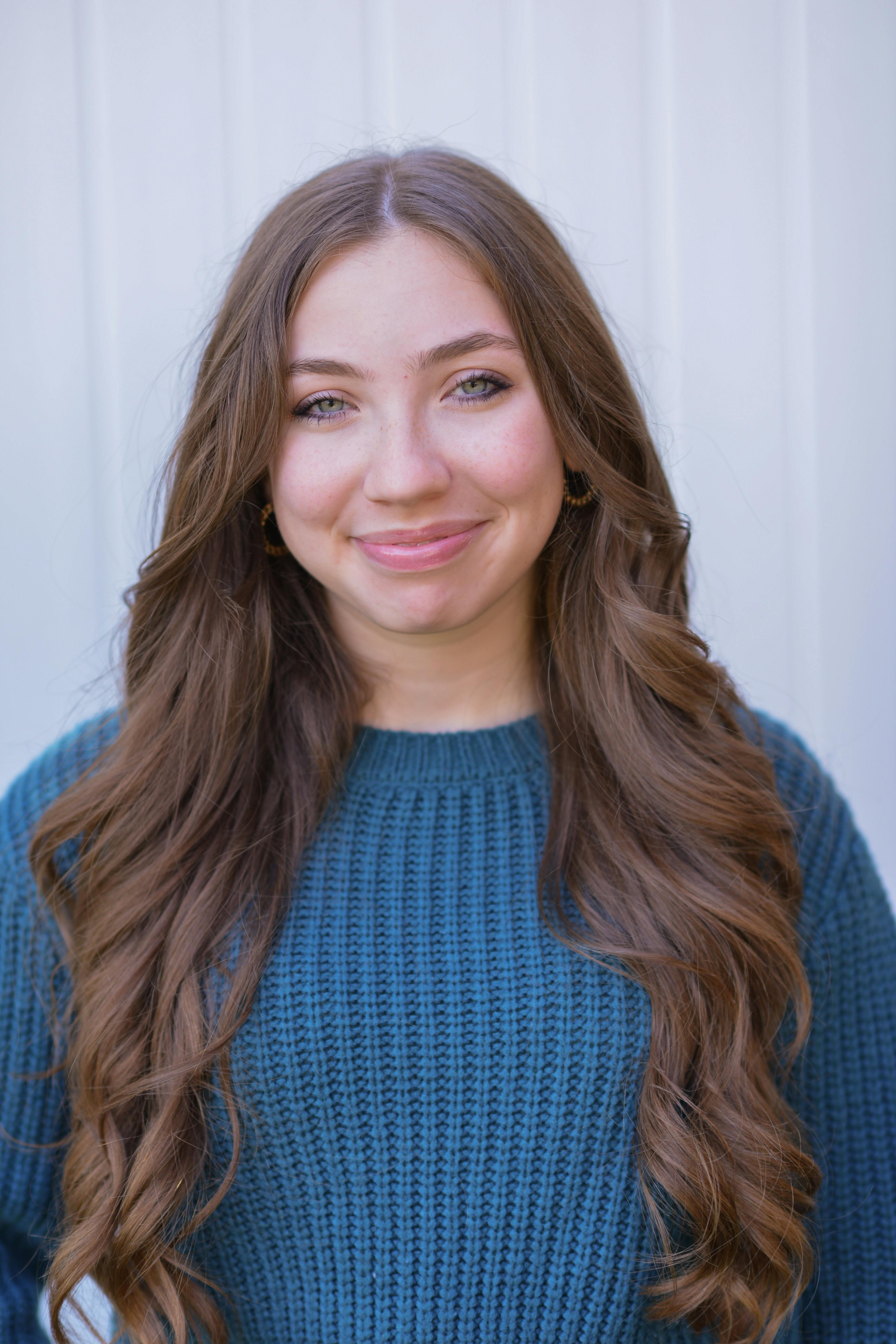 Close-up portrait of a smiling young woman in a blue sweater against a neutral background.