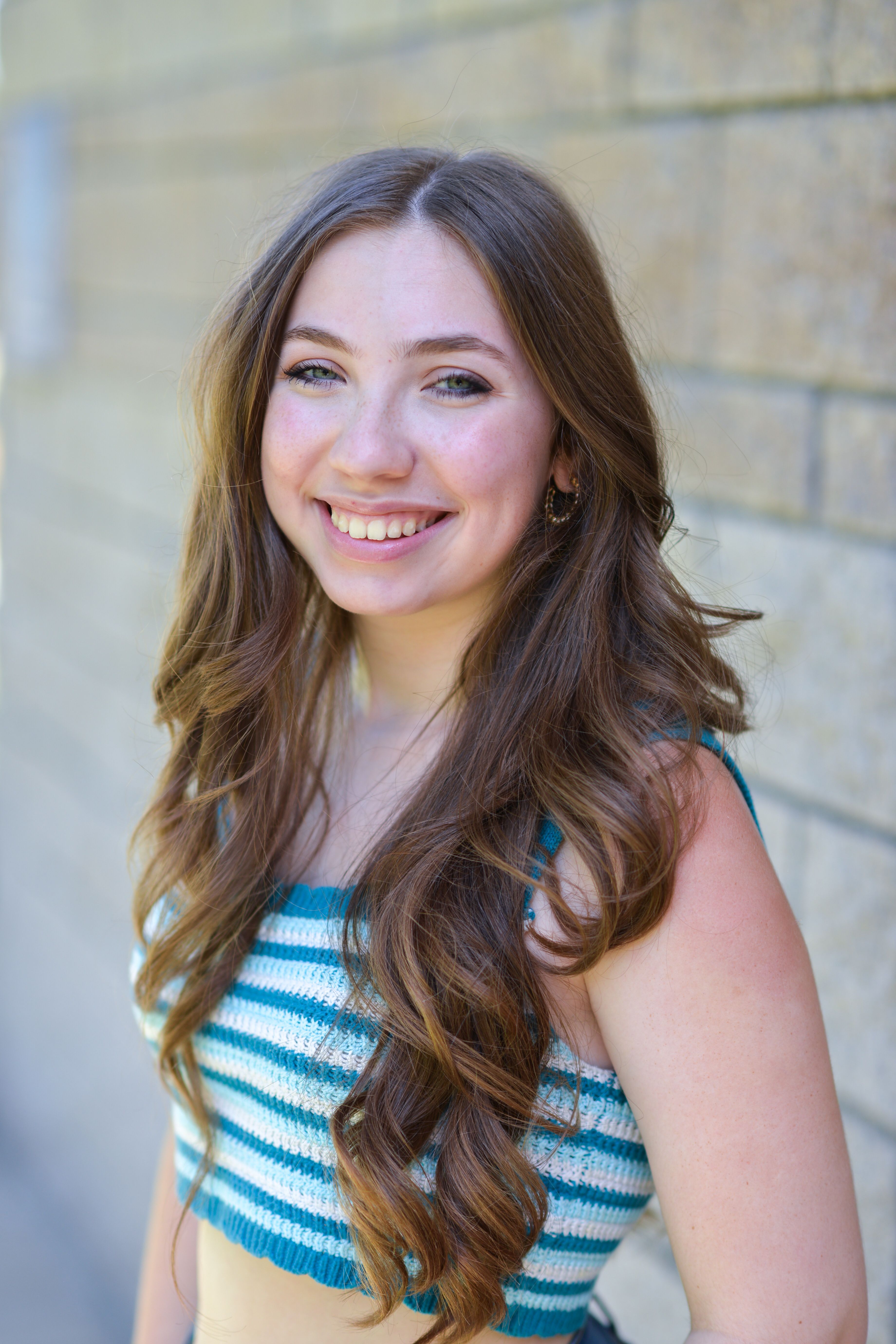 Portrait of a smiling young woman with long brown hair, wearing a striped top, standing outdoors.