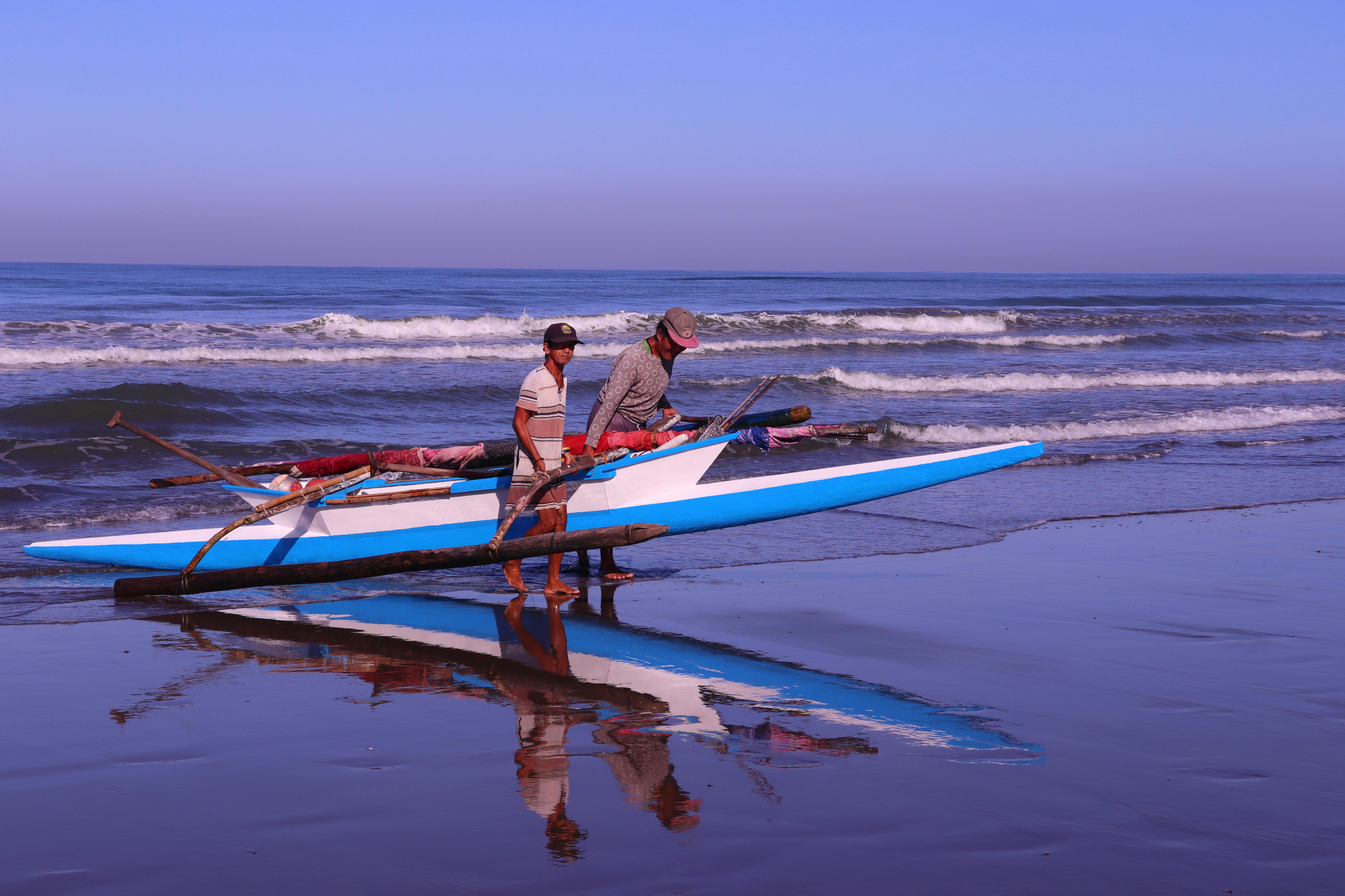 Free Two fishermen prepare a traditional boat on a reflective beach at sunrise. Stock Photo