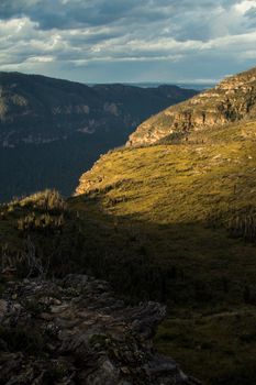 Breathtaking aerial view of a sunlit valley surrounded by majestic mountains and lush greenery under dramatic skies.