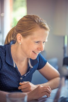 A cheerful woman in a blue shirt smiling while using a laptop indoors, reflecting enjoyment and focus.