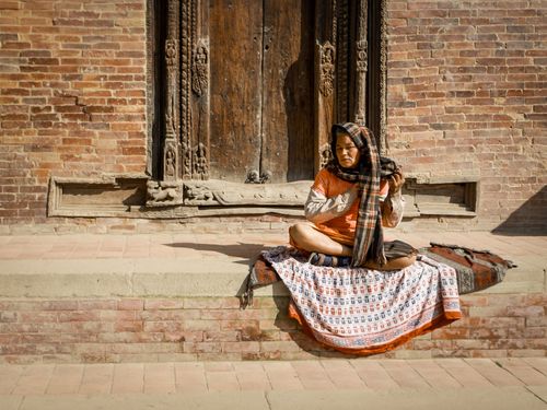 Free Woman in traditional attire sitting by an ornate wooden door against a brick wall, in a street setting. Stock Photo