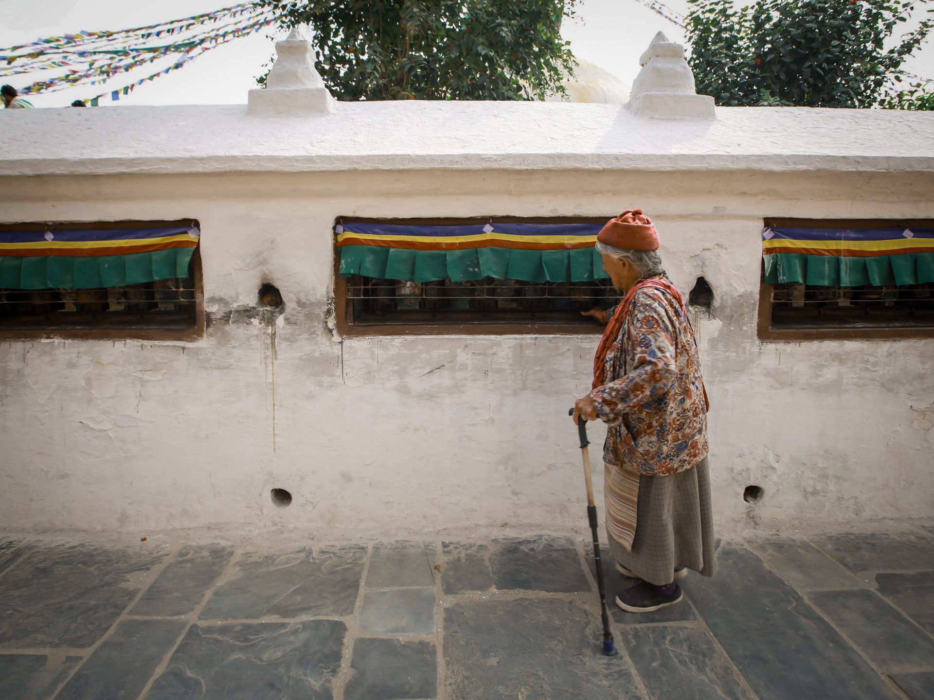 Elderly woman with walking stick by prayer wheels outdoors, capturing cultural essence.