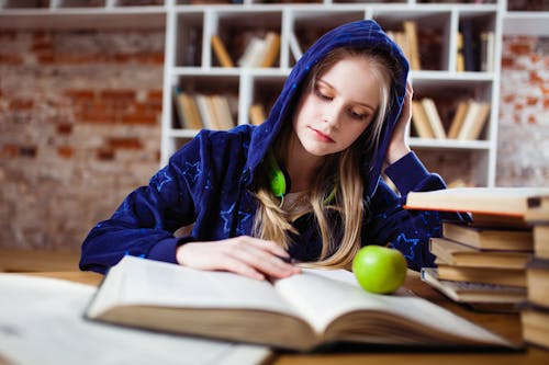 Wanita Mengenakan Jaket Biru Duduk Di Kursi Dekat Meja Membaca Buku