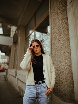 Stylish woman with sunglasses posing against a city wall in a casual outfit, exuding confidence and fashion-forward charm.