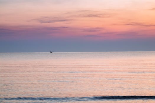 Tranquil sunrise at Hua Hin, Thailand. Soft pink and orange sky reflect on calm waters with a distant boat.