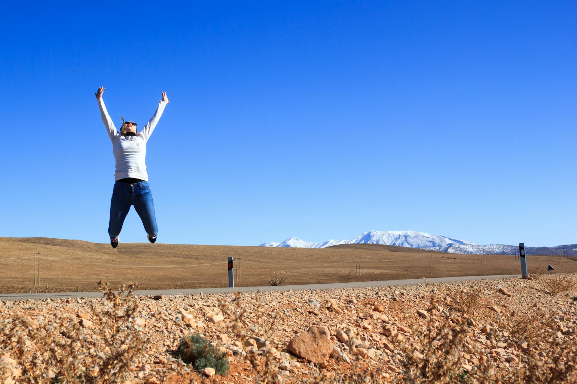 Person jumps joyfully on a road in the arid Moroccan landscape with mountains in the background.