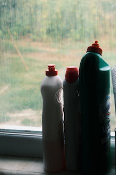 Plastic detergent bottles on a windowsill with a rainy outdoor view, creating a still life scene.