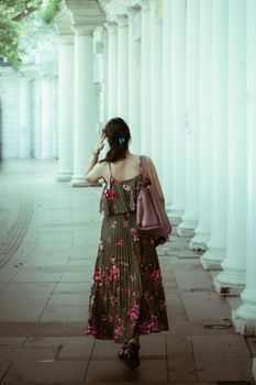 A woman in a floral dress walks down a pillared corridor, carrying a bag. Back view with a cool, calm ambiance.
