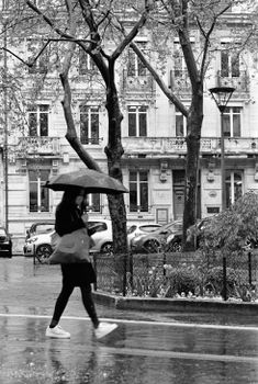 Monochrome scene of a woman walking with an umbrella in a rainy city.