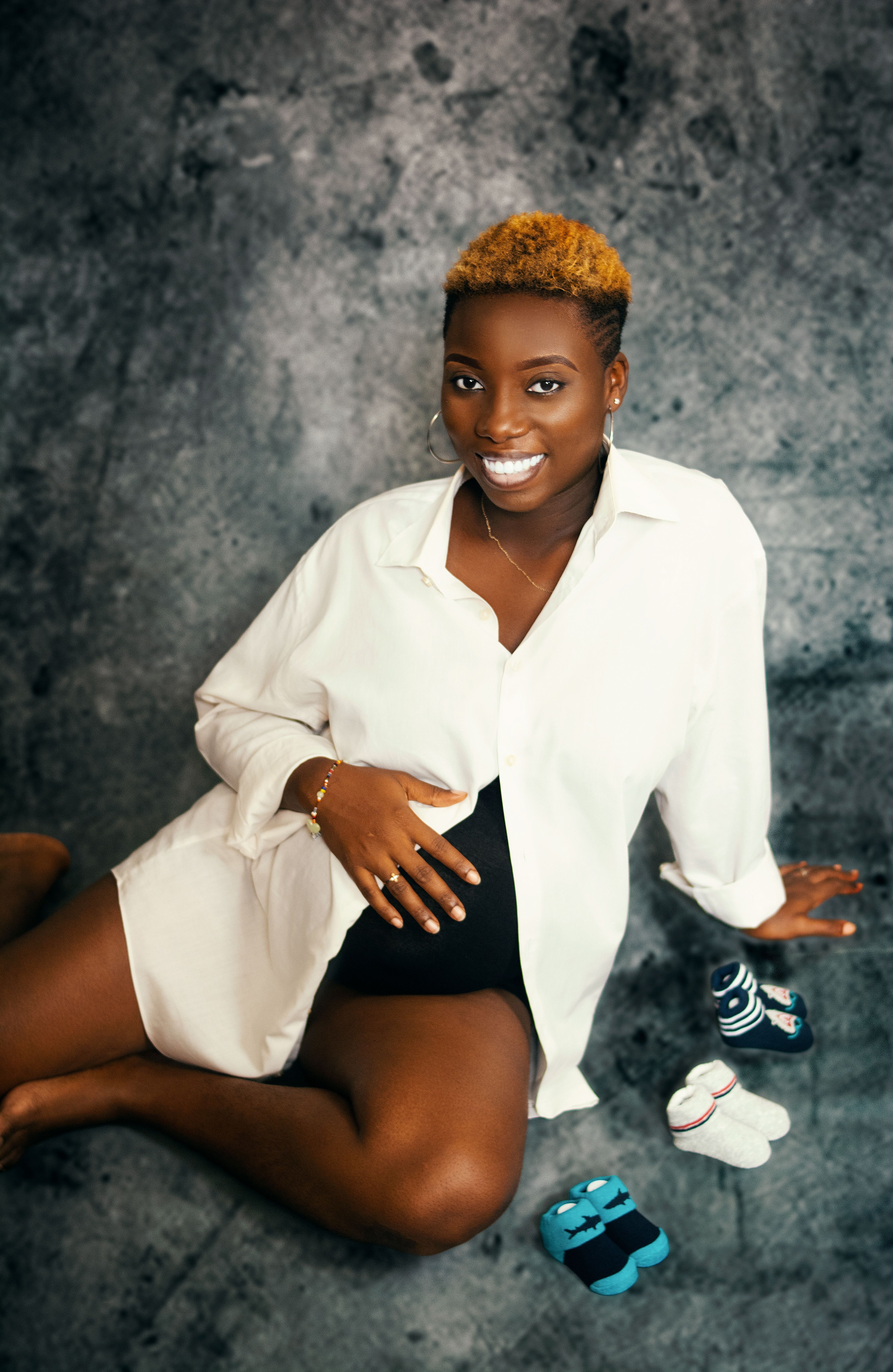 A pregnant Black woman in a white button-up sits on a concrete floor, baby socks beside her, touching her belly.