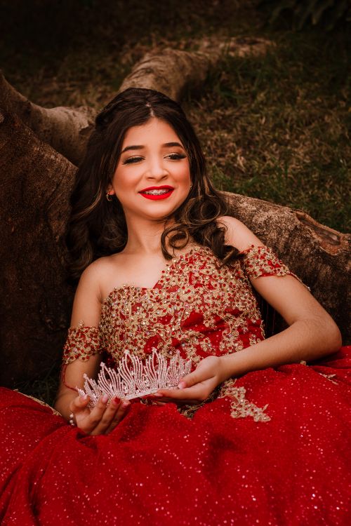 Free Teen girl in red gown smiling and holding tiara outdoors. Elegant portrait with a natural background. Stock Photo