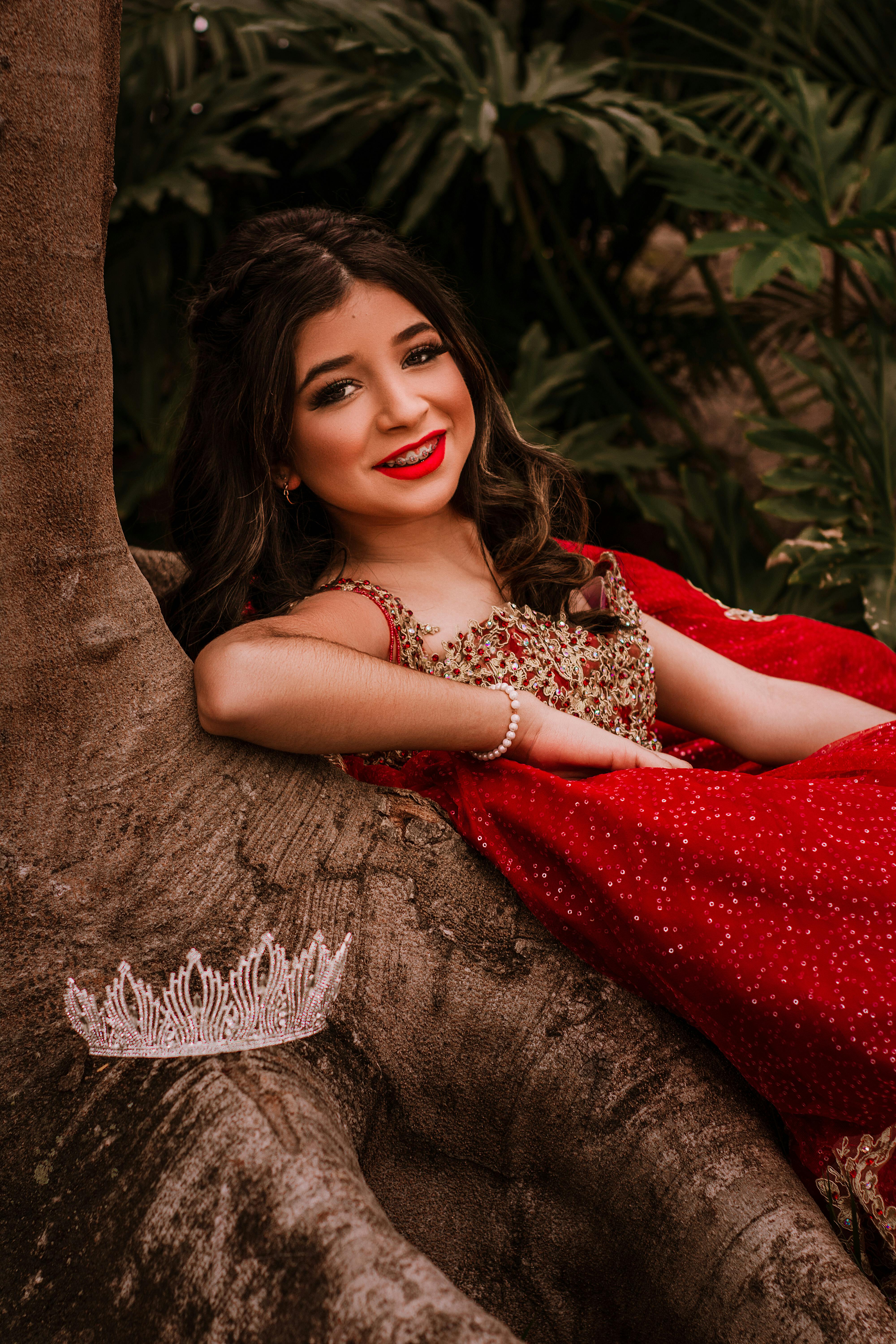 smiling teenage girl in red gown leaning on a tree branch